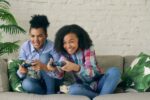 Two smiling girls playing video games on couch.