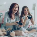 Mother and daughter play video games on a bed.